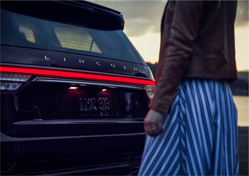 A person is shown near the rear of a 2023 Lincoln Aviator® SUV as the Lincoln Embrace illuminates the rear lights | George Ballentine Lincoln in Greenwood SC