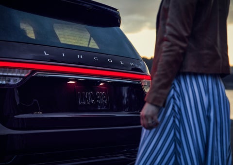 A person is shown near the rear of a 2024 Lincoln Aviator® SUV as the Lincoln Embrace illuminates the rear lights | George Ballentine Lincoln in Greenwood SC